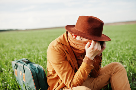 Stylish man in glasses, brown jacket and hat with bag posed on green field.の写真素材