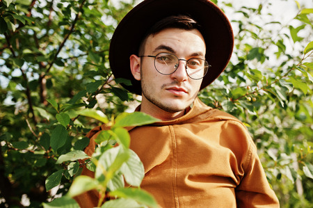 Stylish man in glasses, brown jacket and hat posed on green field.