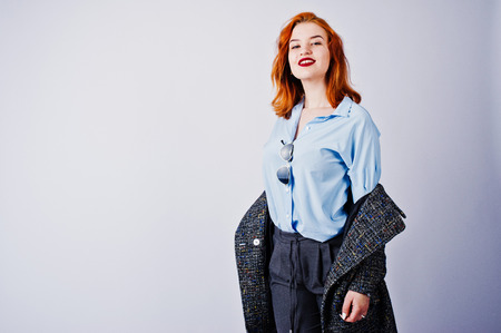 Portrait of a fantastic redheaded girl in blue shirt, grey overcoat posing with sunglasses in the...