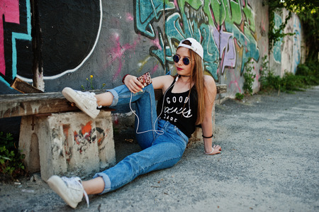 Stylish casual hipster girl in cap, sunglasses and jeans wear, listening music from headphones of...