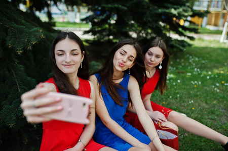 Three teenagers girl in blue and red dresses posed outdoor and making selfie at mobile phone.の写真素材