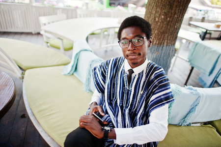 African man in traditional clothes and glasses sitting at outdoor caffe with cell phone.の写真素材