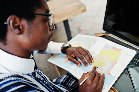 African man in traditional clothes and glasses sitting behind laptop at outdoor caffe and looking on map of Africa and Ghana at his notebook.の写真素材