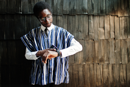 African businessman in traditional clothes and glasses with mobile phone at hand looking at his watches against wooden wall.の写真素材