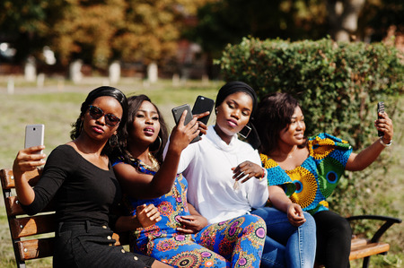 Group of four african american girls sitting on bench outdoor with mobile phones at hands and making selfie.の写真素材