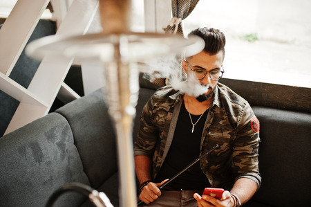 Stylish beard arabian man in glasses and military jacket smoking hookah at street bar. Arab model...