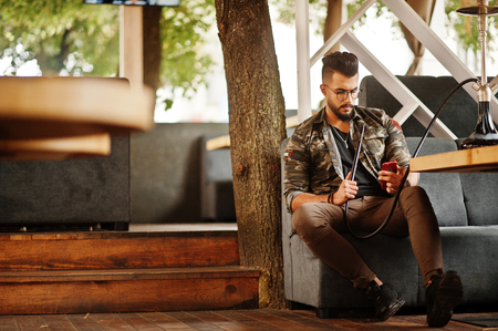 Stylish beard arabian man in glasses and military jacket smoking hookah at street bar. Arab model having rest and looking on phone.の写真素材