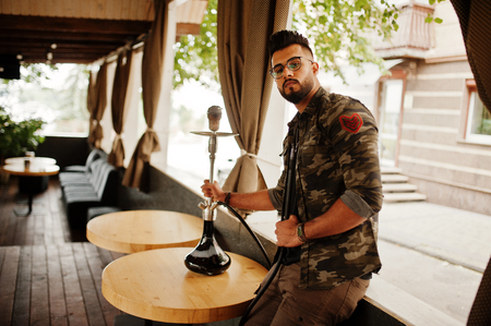 Stylish beard arabian man in glasses and military jacket smoking hookah at street bar. Arab model having rest.の写真素材