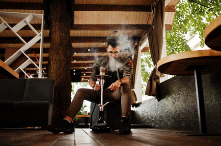 Stylish beard arabian man in glasses and military jacket smoking hookah at street bar. Arab model...