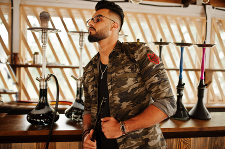 Stylish beard arabian man in glasses and military jacket smoking hookah at street bar. Arab model...