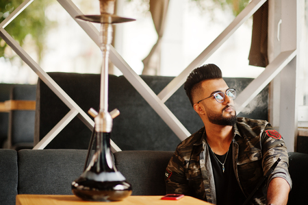 Stylish beard arabian man in glasses and military jacket smoking hookah at street bar. Arab model...