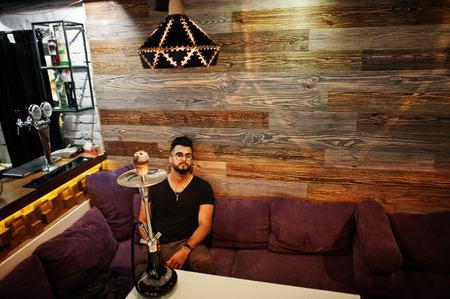 Stylish beard arabian man in glasses and black t-shirt smoking hookah indoor bar. Arab model having...