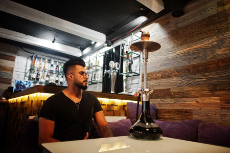 Stylish beard arabian man in glasses and black t-shirt smoking hookah indoor bar. Arab model having...