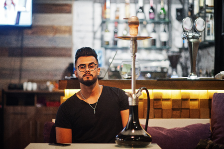 Stylish beard arabian man in glasses and black t-shirt smoking hookah indoor bar. Arab model having rest.の写真素材