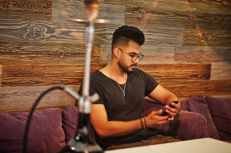 Stylish beard arabian man in glasses and black t-shirt smoking hookah indoor bar. Arab model having...