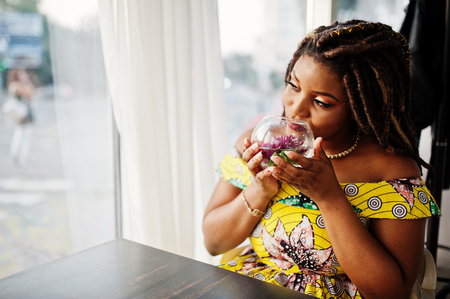 Cute small height african american girl with dreadlocks, wear at coloured yellow dress, sitting at cafe with flower at glass.の写真素材