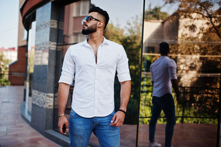 Stylish tall arabian man model in white shirt, jeans and sunglasses posed at street of city. Beard attractive arab guy against modern building.の写真素材