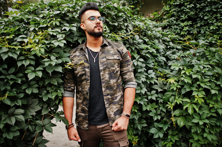 Awesome beautiful tall ararbian beard macho man in glasses and military jacket posed outdoor against arch of green leaves.の写真素材