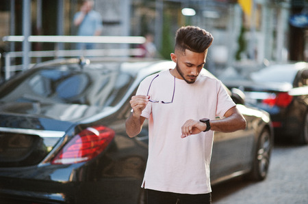 Stylish indian beard man at sunglasses and pink t-shirt against luxury car. India rich model posed outdoor at streets of city and looking at his watches.の写真素材