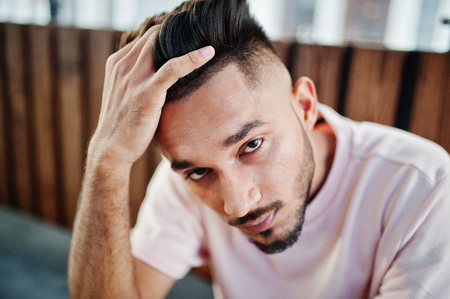 Stylish indian beard man at pink t-shirt sitting against wooden table of cafe. India model posed outdoor. Close up portrait.の写真素材