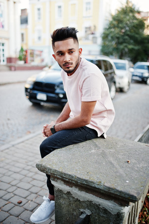 Stylish indian beard man at pink t-shirt sitting against black suv car. India model posed outdoor at streets of city.の写真素材