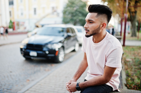 Stylish indian beard man at pink t-shirt sitting against black suv car. India model posed outdoor at streets of city.の写真素材