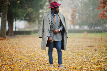 Stylish African American man model in gray coat, jacket tie and red hat posed at foggy weather street at autumn.の写真素材