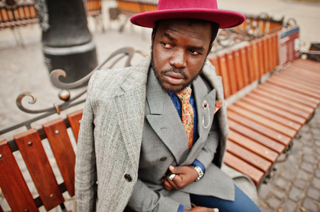 Stylish African American man model in gray coat, jacket tie and red hat posed at bench.の写真素材