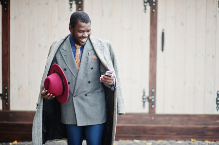 Stylish African American man model in gray coat, jacket tie and red hat posed against wooden cafe and looking at mobile phone.の写真素材