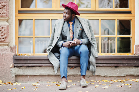 Stylish African American man model in gray coat, jacket tie and red hat sitting against large windows and holding phone at hands.の写真素材