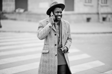 Stylish African American man model in gray coat, jacket tie and red hat walking on crosswalk and speaking on mobile phone. Black and white photo.の写真素材