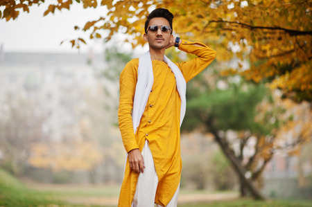Indian stylish man in yellow traditional clothes with white scarf, sunglasses posed outdoor against autumn leaves tree.の写真素材