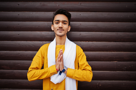 Indian stylish man in yellow traditional clothes with white scarf posed outdoor against wooden background and shows namaste hands sign.の写真素材