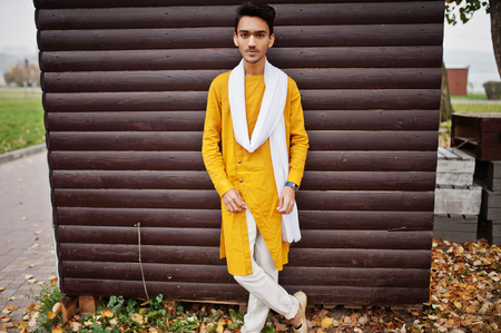 Indian stylish man in yellow traditional clothes with white scarf posed outdoor against wooden background.の写真素材