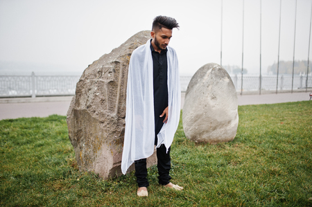 Indian stylish man in black traditional clothes with white scarf posed outdoor against big stones.の写真素材