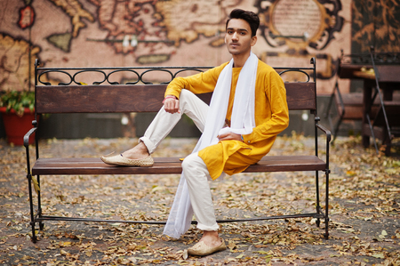 Indian stylish man in yellow traditional clothes with white scarf posed outdoor on bench.の写真素材