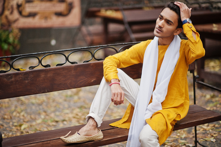 Indian stylish man in yellow traditional clothes with white scarf posed outdoor on bench.の写真素材