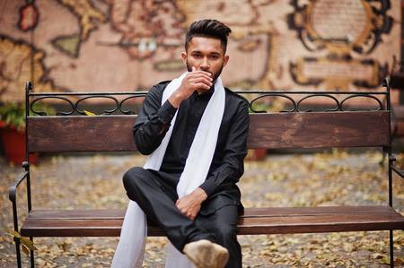 Indian stylish man in black traditional clothes with white scarf posed outdoor sitting on bench.の写真素材
