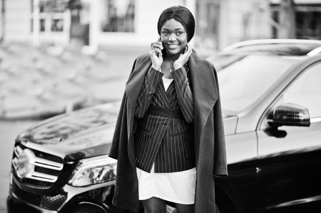 Success stylish african american woman in coat against black business suv car speaking at phone.の写真素材