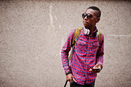 African american man in checkered shirt, sunglasses and earphones with suitcase. Black man traveler against wall holding mobile phone.の写真素材