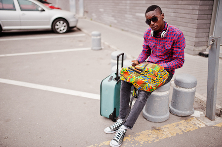 African american man in checkered shirt, sunglasses and earphones with suitcase and backpack. Black man traveler holding mobile phone at hand.の写真素材