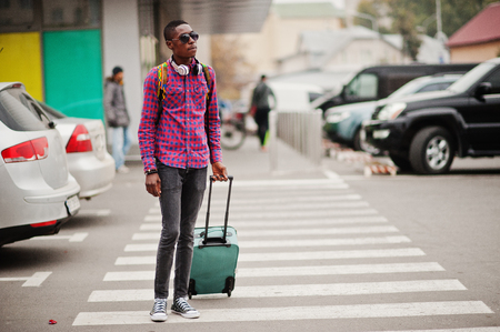 African american man in checkered shirt, sunglasses and earphones with suitcase and backpack. Black man traveler walking on crosswalk.の写真素材