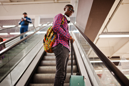 African american man in checkered shirt, sunglasses and jeans with suitcase and backpack. Black man traveler on escalator.の写真素材