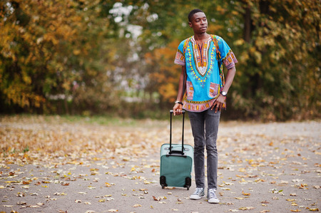 African man in africa traditional shirt on autumn park with backpack and suitcase. Emigrant traveler.の写真素材