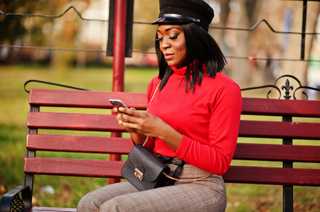 African american fashion girl in newsboy cap, with handbag posed at street, sitting on bench with mobile phone.の写真素材