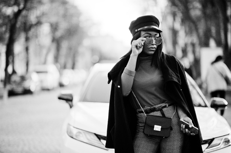 African american fashion girl in coat, newsboy cap and sunglasses posed at street against white business car.の写真素材
