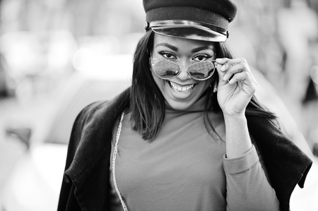 African american fashion girl in coat, newsboy cap and sunglasses posed at street against white business car.の写真素材