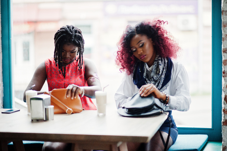 Attractive african american two girls friends sitting at table on cafe and looking something at their handbags.の写真素材