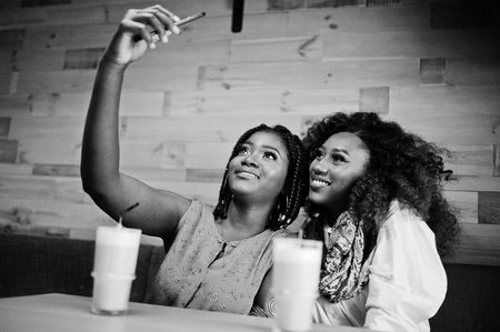 Attractive african american two girls friends sitting at cafe with latte and making selfie on mobile phone.の写真素材