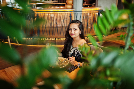 Pretty indian girl in black saree dress posed at restaurant, sitting at table with juice and salad.の写真素材